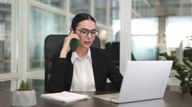 Ofis sandalyesinde oturan ve cep telefonuyla konuşan şık gözlüklü, modern bilgisayar ekranına bakan pozitif iş kadını. İş kavramı ve görevde başarı.