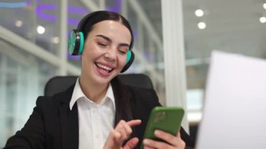 Ofis sandalyesinde oturan ve elinde akıllı telefonla kulaklıkla müzik dinleyen mutlu bir girişimci kadın. Kafkasyalı esmer oda arasında şarkı listesini değiştiriyor ve şarkıların keyfini çıkarıyor..