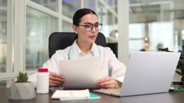 Profesyonel dengeli hemşire masada oturuyor ve online danışmanlık yaparken elinde belgeler tutuyor. Modern bilgisayar kullanan ve klinikteki işyerinde teşhis koyan Kafkas esmer.