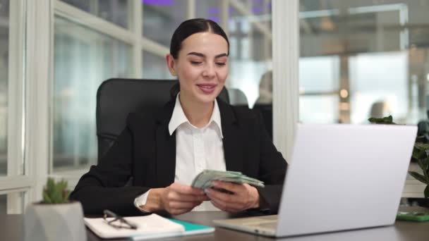 ドル紙幣を手に持っている間 ビデオ通話で話すためにデジタルラップトップを使用する楽観的なマネージャー オンラインインタビューを実施し オフィスで可能な給与を示すプロの女性 — ストック動画
