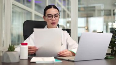 Konsantre kadın terapist masada oturuyor ve hastayla görüntülü konuşma yaparken kablosuz bilgisayar kullanıyor. Elinde kağıt belgeler tutan ve odada bilgi analiz eden güzel yetişkin bir doktor..