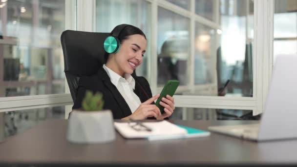 近代的なノートパソコンの近くのテーブルに座り オフィスのヘッドフォンで音楽を聴きながらスマートフォンを保持するリラックスしたビジネスレディ 職場での休憩と趣味のコンセプト — ストック動画