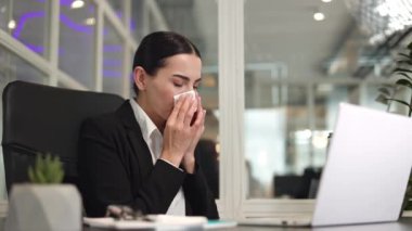 Kablosuz bilgisayarın önünde oturan ve burnunu mendile sümkürürken ekrana bakan kadın ofis çalışanı. Kafkasyalı sağlıksız çalışanın burnu akıyor ve işyerinde kendini kötü hissediyor..