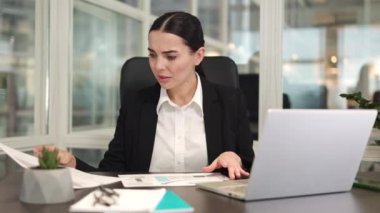Endişeli bir ofis çalışanı iş yerinde oturup içerideki başarısız raporları incelerken modern dizüstü bilgisayar kullanıyor. Saçları örgülü beyaz bir kadın elinde evraklarla ofiste kötü istatistiklere bakıyor..