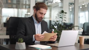 Şaşırmış iş adamı, ofis masasındaki mektupta şok edici haberler okuyor ve zarfı açıyor. Beyaz erkek, kapalı mekanda kişisel dizüstü bilgisayar üzerinde çalışırken işten çıkarılma uyarısı konusunda hayal kırıklığına uğramış..