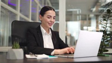 Klasik giysiler içinde gülümseyen kadın ofis çalışanı masada oturuyor ve modern bilgisayarla işini bitiriyor. Başarılı bayan, kapalı alandaki işi bitirdiği için sevinçle sandalyeye yaslanıyor..
