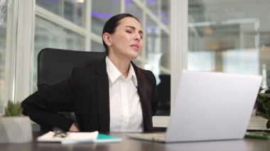 Yorgun bir iş kadını, ciddi sırt ağrısı hissederken masada oturup dijital dizüstü bilgisayarla çalışıyor. Siyah ceketli Kafkasyalı bir çalışan ofis sandalyesinde gerinmeye çalışıyor..