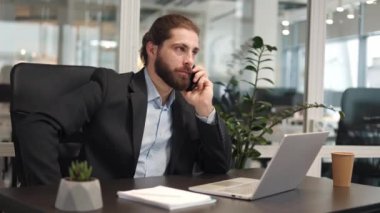 Resmi kostüm giymiş odaklanmış işadamı masadaki kablosuz dizüstü bilgisayarın ve kağıt bardağın yanında modern akıllı telefondan konuşuyor. İş yerindeki iş problemlerini çözmekle meşgul yakışıklı girişimci..