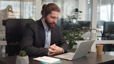 Başarılı sakallı iş adamı takım elbiseli bir patron taşınabilir dizüstü bilgisayarla online toplantı yapıyor ve iş arkadaşlarıyla konuşuyor. Modern ofiste çalışan kulaklıklı beyaz bir erkek..