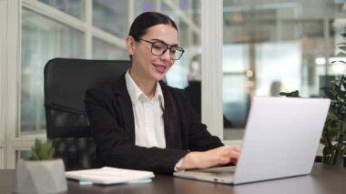 Gülümseyen ofis çalışanının şık gözlükler takıp masada oturması ve klavyede dizüstü bilgisayara yazı yazması. Ofiste internetten bilgi arayan beyaz bir kadın..