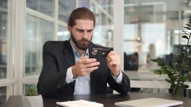 男人坐在手提电脑前的办公桌前 在个人智能手机中输入银行卡的细节时被弄糊涂了 令人惊讶的是 在网上购物和使用现代小玩意的高加索男性 — 图库视频影像