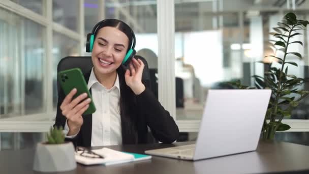 無線ラップトップ近くのテーブルに座り オフィスのヘッドフォンで音楽を楽しんでいる満足の女性従業員 スマートフォンを持っている笑顔の白人女性と仕事で休憩中にリラックス — ストック動画