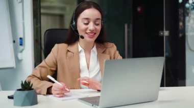 Ofisteki bilgisayardaki video görüşmesi sırasında şirket gelirini arttırmak için kulaklık imzalayan yetkin beyaz iş kadını. Dolabın arka planında bir kadın cihaz üzerinde sohbet ediyor..