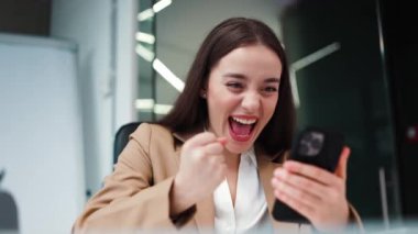 İş kıyafetleri giymiş hırslı bir kadın çalışan iş yerinde başarılı olurken dijital akıllı telefona bakıyor. Zaferi kutlarken masada oturan mutlu esmer kadın..