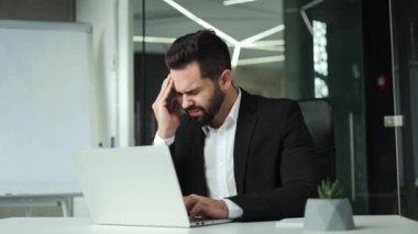 Hayal kırıklığına uğramış işadamı, portatif dizüstü bilgisayarın önünde otururken ağrı hissettiği için kafaya dokunuyor. Ofiste çalışmak için modern aletler kullanan yorgun Kafkas çalışanı..