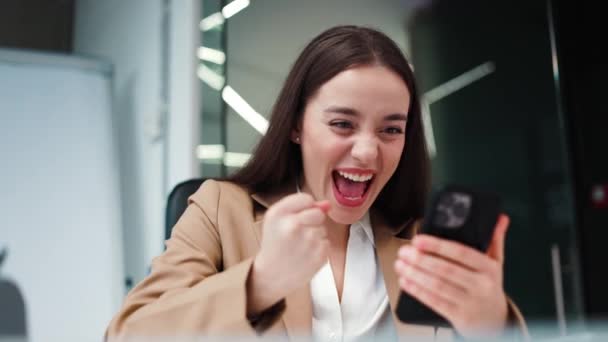 職場での成功を楽しみながら デジタルスマートフォンを見ているビジネス服を着た野心的な女性社員 オフィスのジェスチャーで勝利を祝いながらテーブルに座っているハッピーブルネット — ストック動画