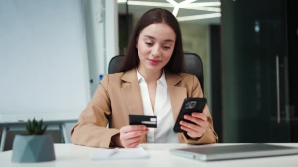 デジタルスマートフォンを手に持っている笑顔の女性マネージャーとブラッククレジットカードのデータを比較します テーブルに座って 作業時間中にオンラインで購入する正式な衣装のコーカサス従業員 — ストック動画