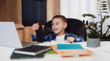 Kafkasyalı çocuk cep telefonunu elinde tutarken portatif bilgisayar ve kitaplarla masanın başında oturuyor. Çocuk, e-öğrenme ya da eğlence amaçlı farklı çevrimiçi etkinlikler için dijital aygıt kullanıyor..