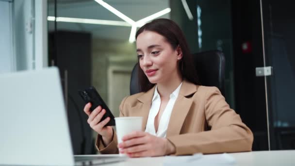 穿着米色西服的积极的女商人拿着一杯咖啡 与现代计算机坐在桌旁 白种人漂亮的经理在办公室休息时看着无线电话 — 图库视频影像