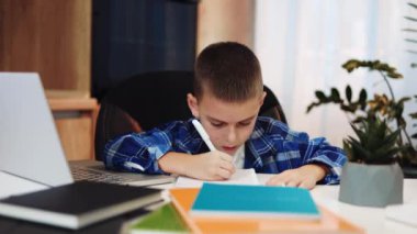 İnternetteki dersler sırasında kablosuz bilgisayarda günlük giysiler içinde kitap yazan gülümseyen çocuk. Akıllı beyaz çocuk kameraya bakıyor ve özel odadaki masada eğitime sevgisini ifade ediyor..