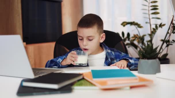 チェックされたシャツの白人子供は 現代のラップトップとデスクの前にオンラインレッスンの間でミルクのガラスを持っています 自宅からオンラインクラスを始める前に健康的なスナックを持っている19人の子供 — ストック動画