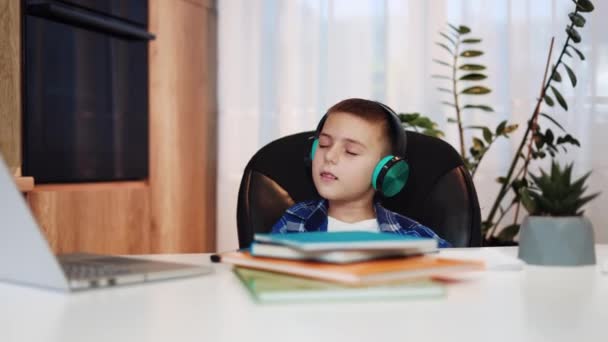 活跃的孩子在无线耳机里听流行音乐 坐在课桌前摇头打闹 有趣的男孩从枯燥无味的家庭作业中放松 一个人在客厅里玩得很开心 — 图库视频影像