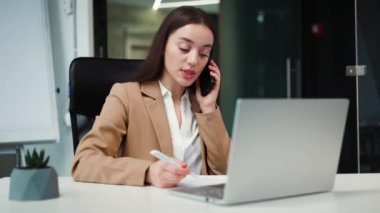 Ciddi yüz ifadesi olan kendine güvenen bir iş kadını modern iş merkezindeki telefon görüşmeleri sırasında not alıyor. Meşgul bayan girişimci partnerle konuşuyor ve dizüstü bilgisayar üzerinde çalışıyor..