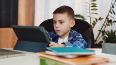Mavi kareli tişörtlü akıllı erkek çocuk, şık oturma odasında otururken internet ve tablet kullanıyor. Çevrimiçi eğitim ve modern teknoloji kavramı.