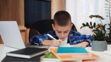 Ev içi iç mekanlardaki çevrimiçi dersler sırasında not defterine not almakla meşgul. Uzaktan öğretmenlerle iletişim kurmak için modern dizüstü bilgisayar kullanan sıradan giyinmiş beyaz bir erkek çocuk..