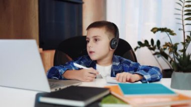 Kulaklığı olan zeki bir okul çocuğu ev içi öğretmeniyle bilgisayarında video görüşmesi yapıyor. Kendine güvenen beyaz erkek çocuk dikkatle yeni bilgileri ve yazıları dinliyor..