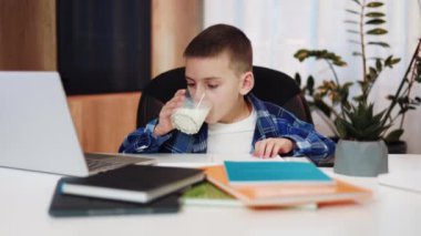 Zeki çocuk, online derste renkli çalışma kitaplarıyla bir bardak süt içip dizüstü bilgisayar başında yazı yazıyor. Çocuklar için başarılı bir eğitim için sağlıklı ve besleyici atıştırmalıkların bulunması.