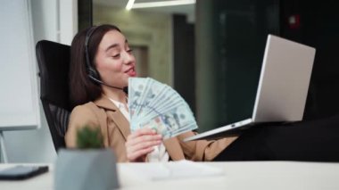 Kulaklık takmış neşeli bir kadın, elinde dolar banknotlarının hayranı ve taşınabilir bilgisayarda online sohbet ediyor. Olumlu kadın yönetici modern aleti elinde tutuyor ve ofis koltuğunun arkasına yaslanıyor..