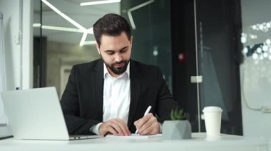 Kafkasyalı girişimci, taşınabilir dizüstü bilgisayarda yazı yazıyor ve ofisteki bir kağıt parçasına bilgi not alıyor. İşyerindeki not defterine iş projesi hakkında bilgi yazan kendinden emin bir adam..