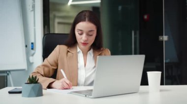 Kafkasyalı esmer, kağıt notlar üzerine bilgi yazarken modern dizüstü bilgisayarla beyaz masada oturuyor ve iş merkezinde bir fincan kahve içiyor. Ofis kavramı, çevrimiçi çalışma ve aygıtlar.