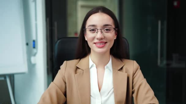 Portrait of attractive caucasian woman wearing trendy glasses sitting in office chair and sincerely smiling. Successful lady with dark hair looking at camera and showing confidence at workplace.
