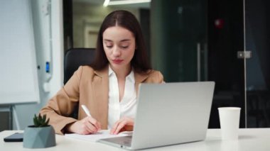 Kafkasyalı girişimci, taşınabilir dizüstü bilgisayarda yazı yazıyor ve ofisteki bir kağıt parçasına bilgi not alıyor. İşyerindeki not defterine iş projesi hakkında bilgi yazan kendinden emin bir kadın..