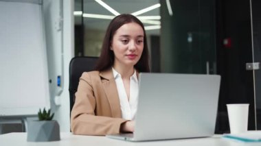 Güzel yüz hatları olan beyaz kadın portresi geniş, aydınlık iş merkezinde dizüstü bilgisayarla masada oturuyor. Çekici esmer kadın iş elbisesi giymiş kameraya bakıyor..