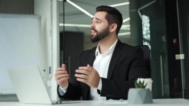 Başarılı sakallı girişimci internetteki kablosuz bilgisayardaki toplantı sırasında el sallıyor. Beyaz, siyah takım elbiseli yakışıklı adam ofis masasında oturuyor ve uzaktan iletişimin tadını çıkarıyor..