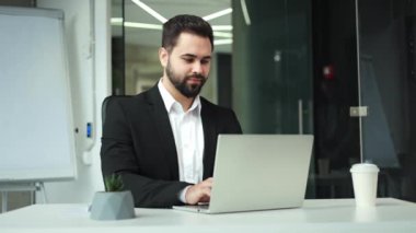 İş ortamının arka planında otururken el hareketi gösteren bir iş adamı portresi. Gülümseyen beyaz erkek modern dizüstü bilgisayar kullanıyor. Onaylayıcı ifadeler veriyor ve doğrudan bakıyor..