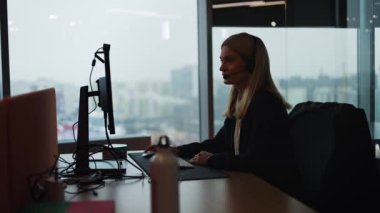 Ofisten internet üzerinden panoramik pencere teknik destekli internet iletişim mikrofonlu bir kulaklıkla yardım masasında çalışan yetişkin müşteri destek operatörü asistanının silueti görünümü.