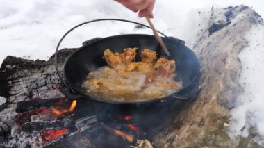 Kafkasyalı adamın, açık havada yanan odunların üzerinde ahşap spatulayla metal tencerede kızartılmış et pişirmesine yakın durun. Vahşi doğada yemek hazırlamak ve kamp ateşinden korunmak..