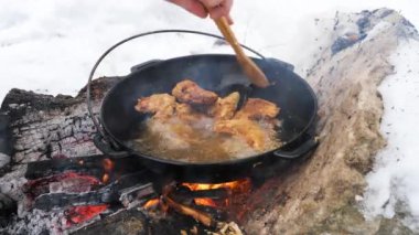 Kışın kar yağarken, metal tencerede ahşap spatulayla tavuk kanatları karıştıran bir adamın elini yakın çekim. Vahşi doğada yemek pişirme ve soğukta doğayla baş başa zaman geçirme..