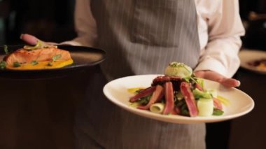 Zarif önlük giymiş profesyonel bayan garson. İki elinde de restoran misafirleri için süslü tabaklar var. Mükemmel servis ve güzel yemek pişirme başyapıtları sunumu.