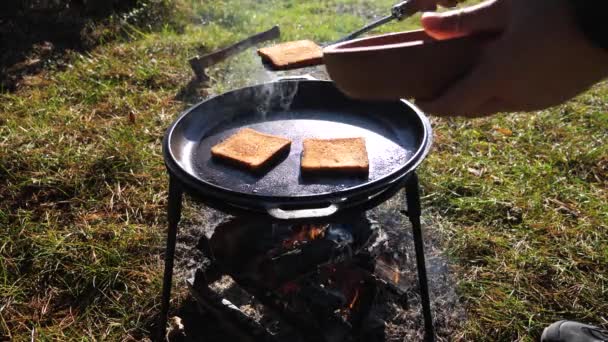 ピクニックでスパチュラと料理しながらアイアンフライパンからパンのスライスを拾う男性の手の近く オープン火のバーベキューグリルで揚げたホットとクランチトーストの準備 — ストック動画