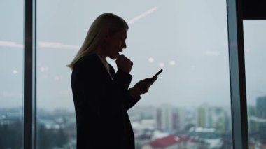 Genç mutlu kadın ofisteki akıllı telefondan iyi haberler okuyor. Kadın işçi cep telefonunu yanında tutuyor, panoramik pencereli siluetler, zaferi kutlayan kadın, online piyango kumarı kazanan..