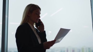 Modern bir cam ofiste cep telefonuyla konuşan bir iş kadınının silüeti gökdelen penceresinin arka planında. Başarılı, kendine güvenen bir iş kadını elinde belge tutuyor..