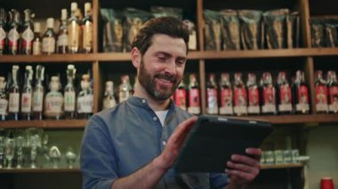 Gülümseyen işletme sahibi kendi kafesinde duruyor ve dijital tablette özel uygulama kullanıyor. Koyu saçlı ve sakallı çekici bir adam modern aletler kullanarak çalışanları kontrol ediyor..