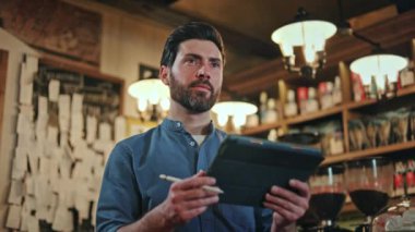 Dost canlısı, mavi üniformalı beyaz adam barda çalışırken dijital tablete bilgi ekliyor. Stylus kalem elinde uzman personel modern işyerindeki misafirden sipariş alıyor..