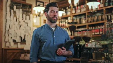 İş yerinde elinde dijital tabletle poz veren mavi gömlekli gülümseyen restoran müdürünün portresi. Koyu saçlı ve sakallı beyaz adam modern uygulamalarla müşteri hizmetlerini izliyor..