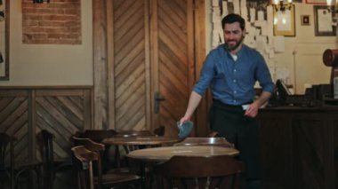 Kafede masaları temizlerken gülümseyen ve kameraya bakan çekici bir adam. Mavi gömlekli memnun barmen eski bistronun mobilyalarını siliyor ve sabah vardiyasına hazırlanıyor..